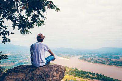 Rear view of man sitting on cliff against sky