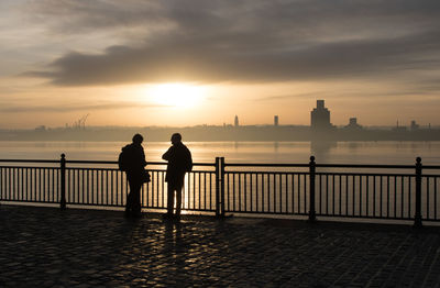 Men standing on railing by sea against sky during sunset