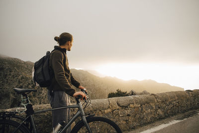 Side view of young man looking at mountains while standing with bicycle on road