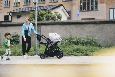 Mother walking with son while pushing baby stroller on sidewalk at city