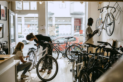 Female owner repairing bicycle of senior customer while colleague working on bike at workshop