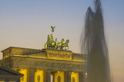 Low angle view of fountain splashing by brandenburg gate against sky during sunset