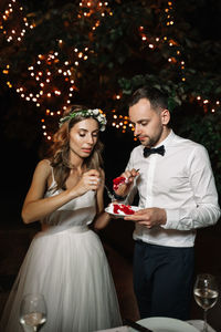 Bride and bridegroom eating dessert while standing against illuminated tree at night