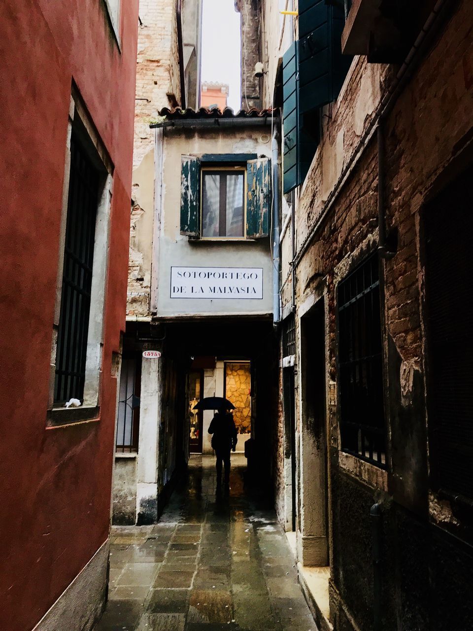 REAR VIEW OF MAN WALKING IN ALLEY AMIDST BUILDINGS