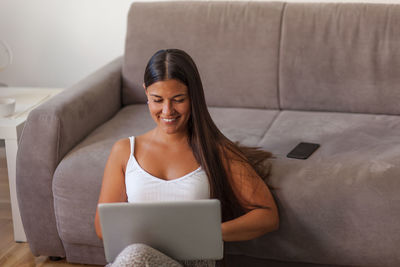 Smiling woman using laptop while sitting by sofa at home