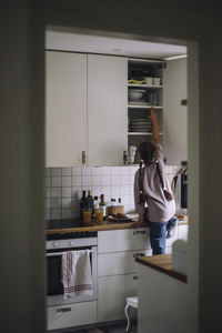 Rear view of girl reaching at cabinet in kitchen at home