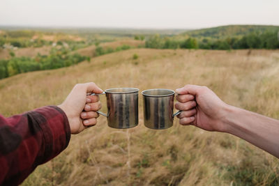 Cropped hands toasting cups over land against sky