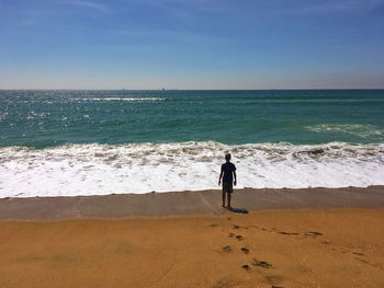 Rear view full length of boy standing at beach against sky on sunny day