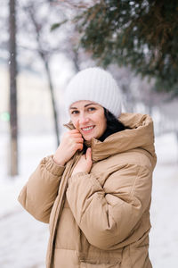 A happy woman stands in a coat and warms her hands. it stands in the city in winter under the snow