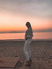 Full length of young woman in white and sunglasses standing on beach at sunset