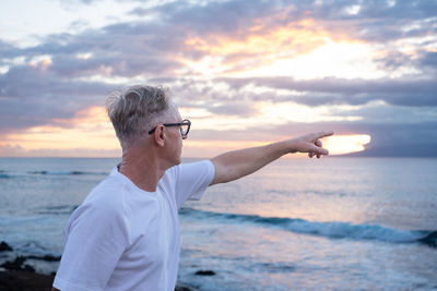 Portrait of young man with arms raised standing at beach against sky during sunset