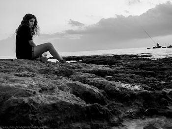 Young woman sitting on rocky seashore against sky