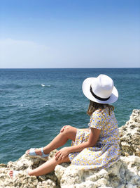 Rear view of woman sitting on rock by sea against clear sky