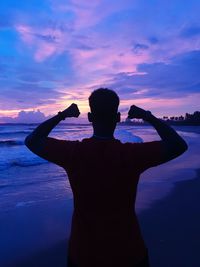 Rear view of man flexing muscles at beach against sky during sunset