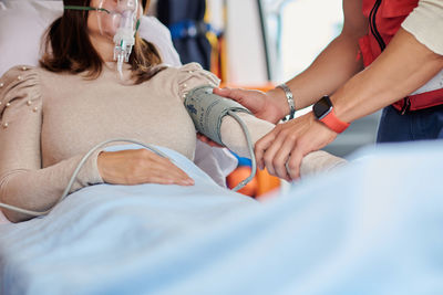 Crop male paramedic putting tensiometer on arm on patient in ventilator mask lying on medical stretcher in ambulance car