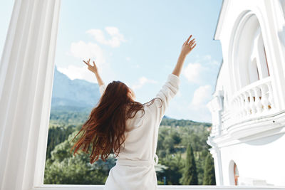 Rear view of woman with arms raised standing by window