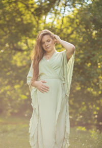 Young pregnant woman stands in park,looks at belly, green trees on background. brown haired female