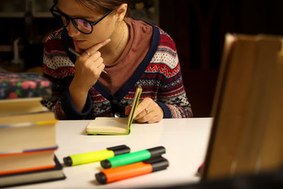 Young woman in glasses study at night, thinks. millennial student holding light green notebook. 