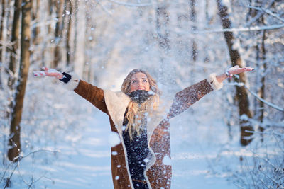 Happy woman throwing snow while standing in forest