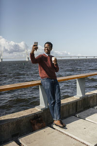 Young man taking smartphone selfie at the sea, holding take out coffee