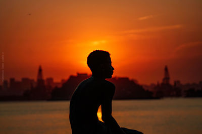 Silhouette man sitting at beach against orange sky
