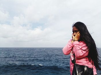 Thoughtful young woman standing at beach against cloudy sky