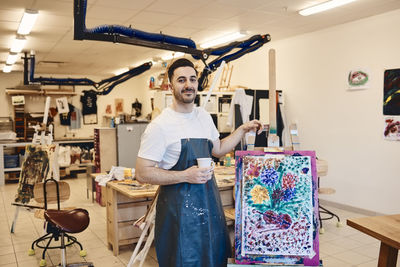 Portrait of confident male artist standing by easel with painting in art class