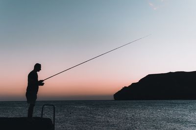 Silhouette man fishing by sea during sunset