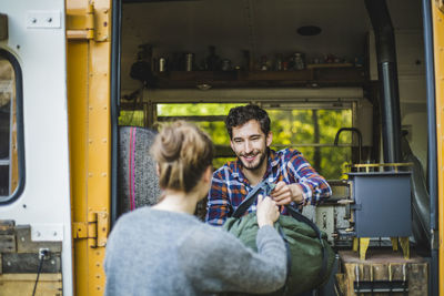 Smiling man giving luggage to woman for unloading from caravan during camping