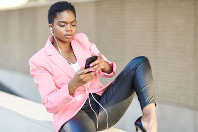 Full length of young woman listening music while sitting on retaining wall outdoors