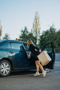 Shopping, sale, happy young stylish blonde woman with shopping bags opening car after shopping. 