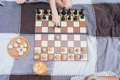 Cropped unrecognizable kids playing chess while sitting on textile with jug of drink and biscuits on lawn