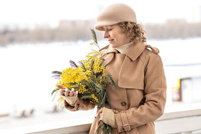 Portrait of a young woman in a beige coat and hat with a bouquet of mimosa in her hands. spring