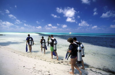 Rear view of scuba divers wearing aqualung walking on sand at beach against sky