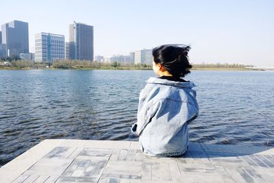 Rear view of woman sitting on pier over river in city