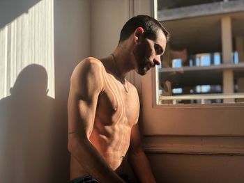 Shirtless young man standing by window at home