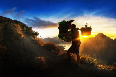 Silhouette man carrying plastic bottles on mountain against sky during sunset