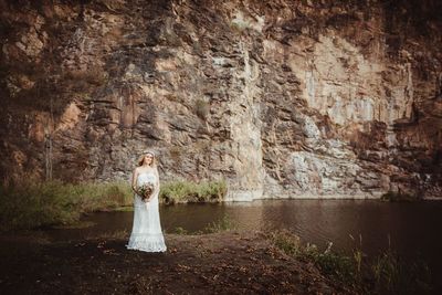 Portrait of woman wearing wedding dress holding bouquet standing by water against rock