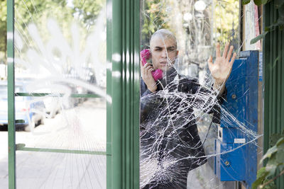 Portrait of bald man using telephone booth seen through shattered glass9