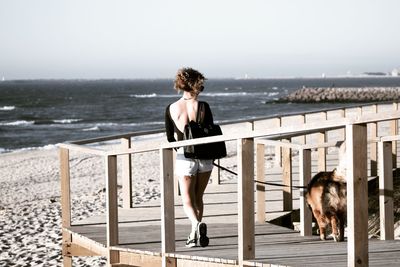 Rear view of young woman walking with dog on boardwalk at beach