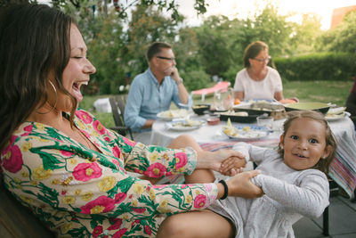 Cheerful mother holding hands of daughter while sitting on chair during garden party