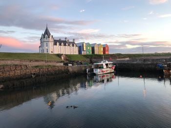 Boats in river by buildings against sky during sunset