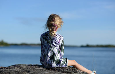 Rear view of girl sitting on rock by sea against sky