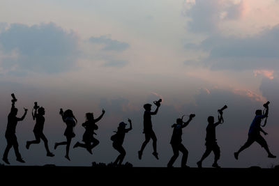 Silhouette people jumping while holding camera against sky during sunset
