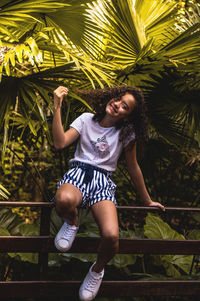 Full length portrait of girl smiling while sitting on railing against palm trees