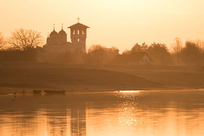 Lighthouse by lake and buildings against sky during sunset