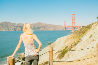 Rear view of woman standing at sea shore by golden gate bridge against clear blue sky