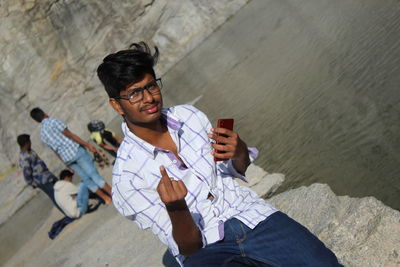 Portrait smiling young man with smart phone showing obscene gesture by lake