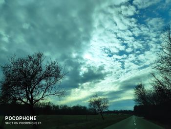 Road by silhouette bare trees on field against sky