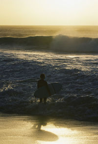 Rear view of surfer walking at beach against sky during sunset